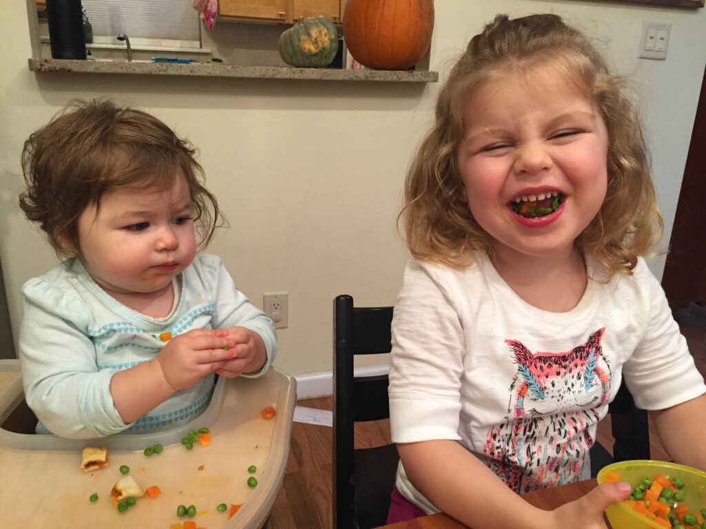 Young child trying solids next to older sister, feeding becomes increasingly solid